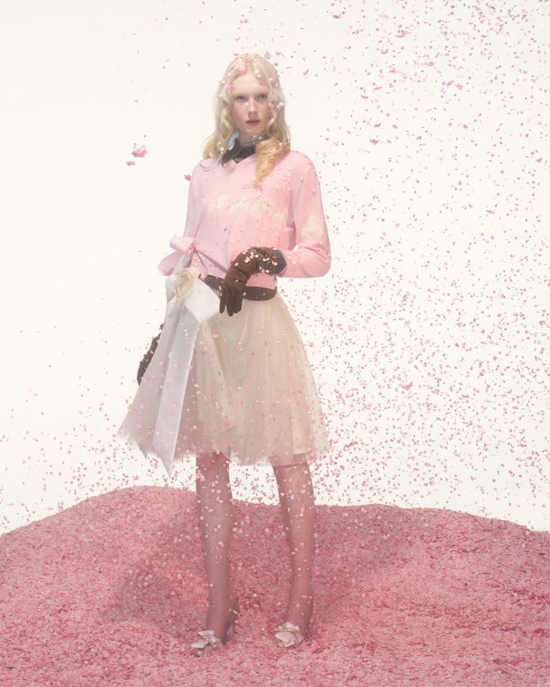 A woman stands in a pink set wearing a pink top, cream-colored skirt, brown gloves, and heels with falling pink confetti.