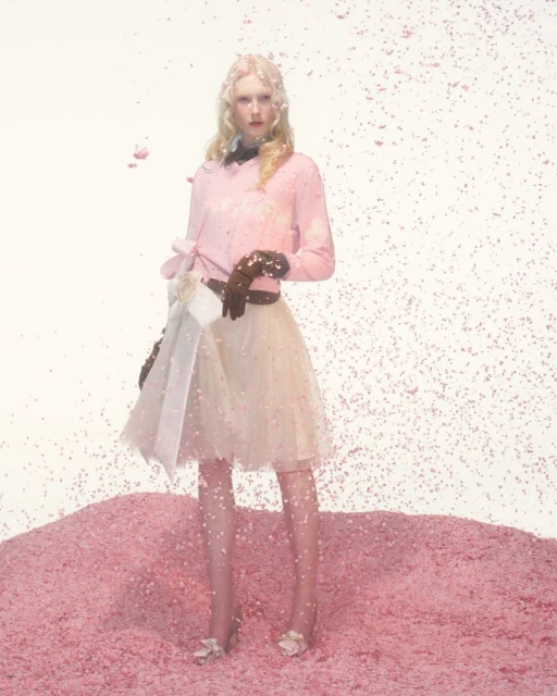 A woman stands in a pink set wearing a pink top, cream-colored skirt, brown gloves, and heels with falling pink confetti.