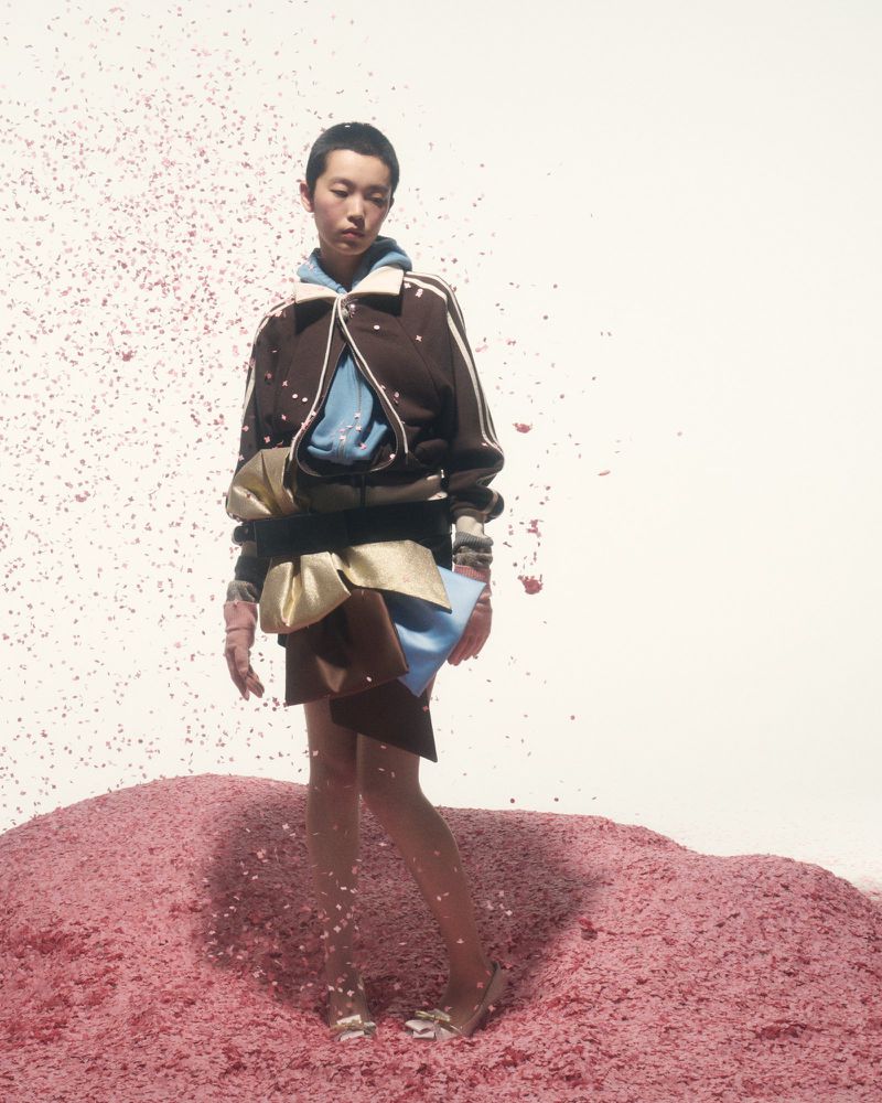 Fashion model posing in a brown bomber jacket, short skirt, and heels, standing on a pile of pink rubble against a white backdrop.
