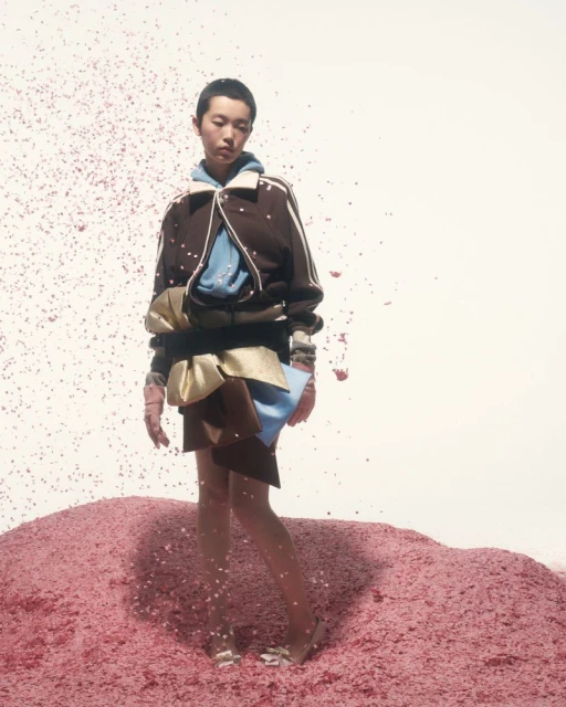 Fashion model posing in a brown bomber jacket, short skirt, and heels, standing on a pile of pink rubble against a white backdrop.