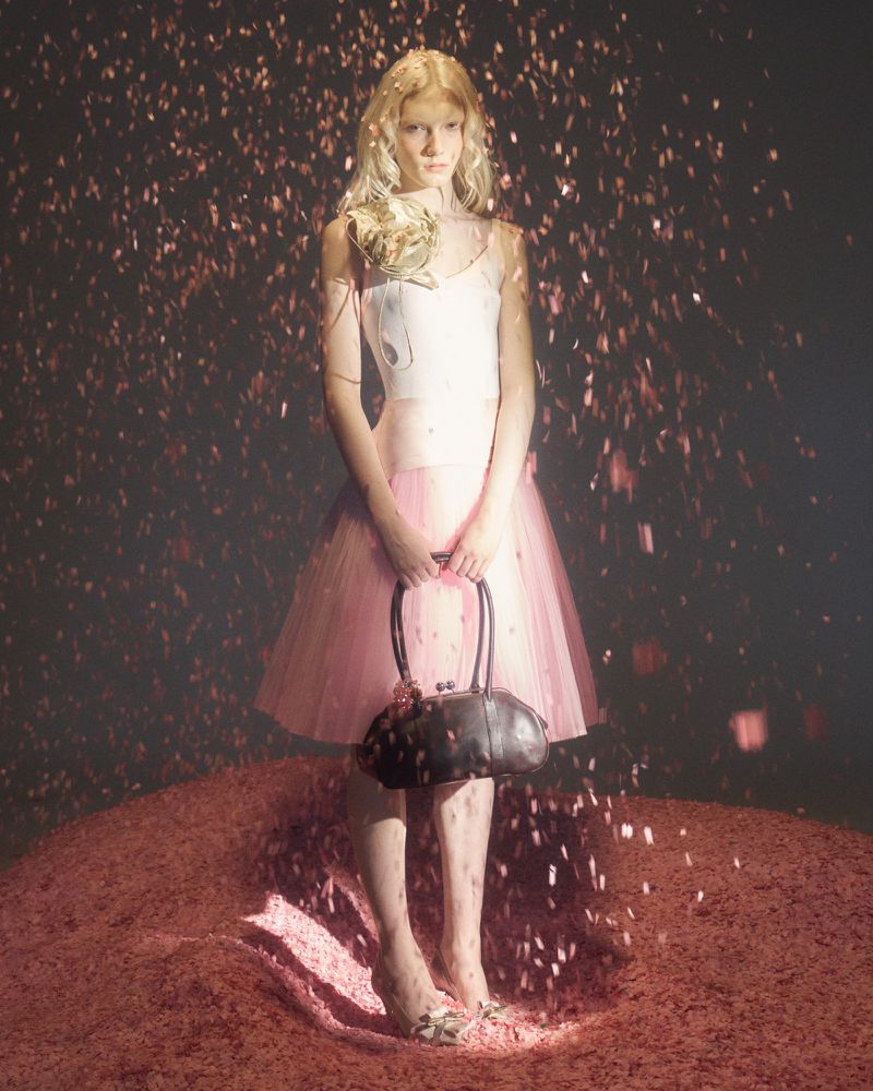 A young fair-skinned woman stands in a pink dress, holding a dark handbag, on a surface covered with small pink pieces, with confetti falling.