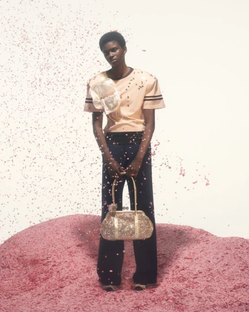 A Black female fashion model poses in a studio surrounded by pink confetti, wearing a peach top and black pants, holding a sequined bag.