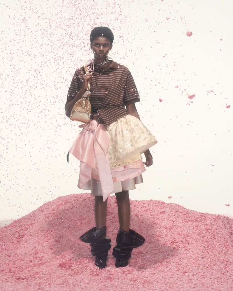 Fashion shot of a woman in a brown striped shirt, tiered skirt, holding a purse, standing on a pink textured surface, against a white backdrop.