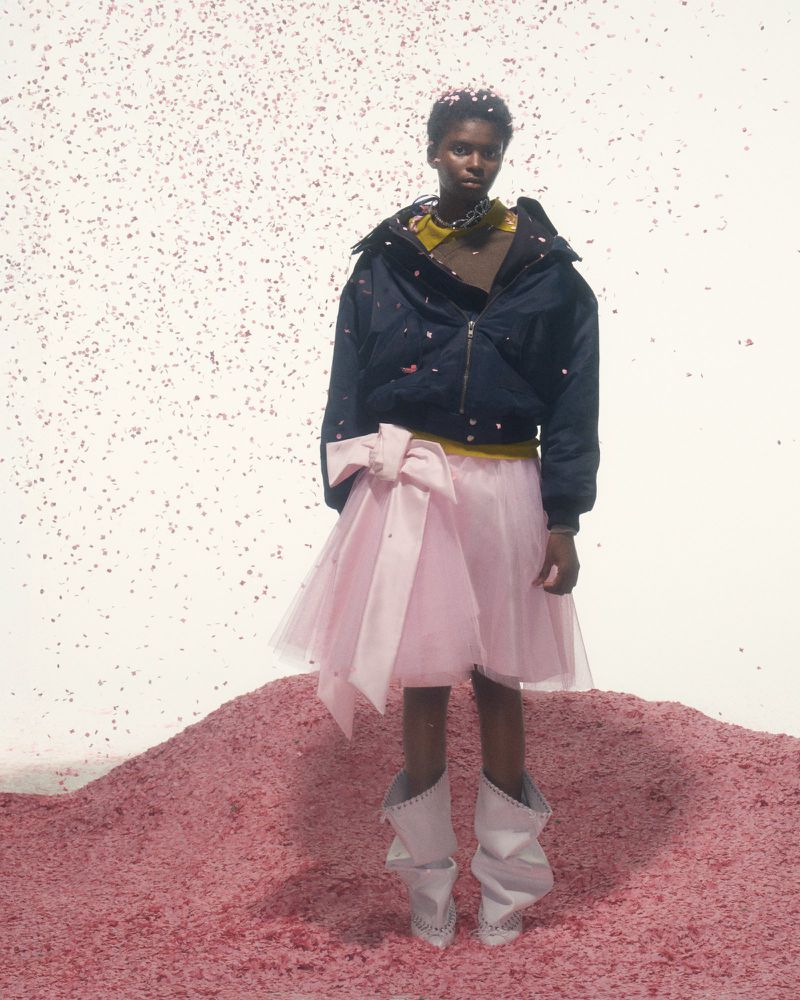 Model in a navy puffer jacket and pink tulle skirt stands on a pink confetti-covered surface in a studio setting, confetti falling.