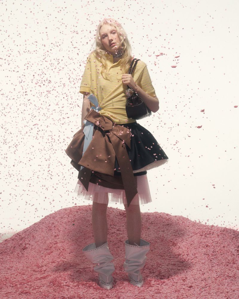 Young woman in yellow top and layered skirt stands on pink confetti hill with matching falling confetti in a fashion editorial shot.