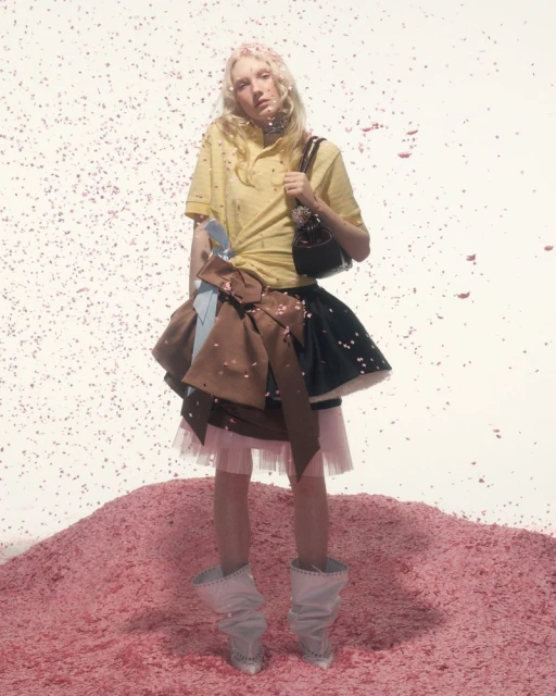 Young woman in yellow top and layered skirt stands on pink confetti hill with matching falling confetti in a fashion editorial shot.