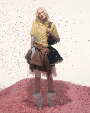 Young woman in yellow top and layered skirt stands on pink confetti hill with matching falling confetti in a fashion editorial shot.