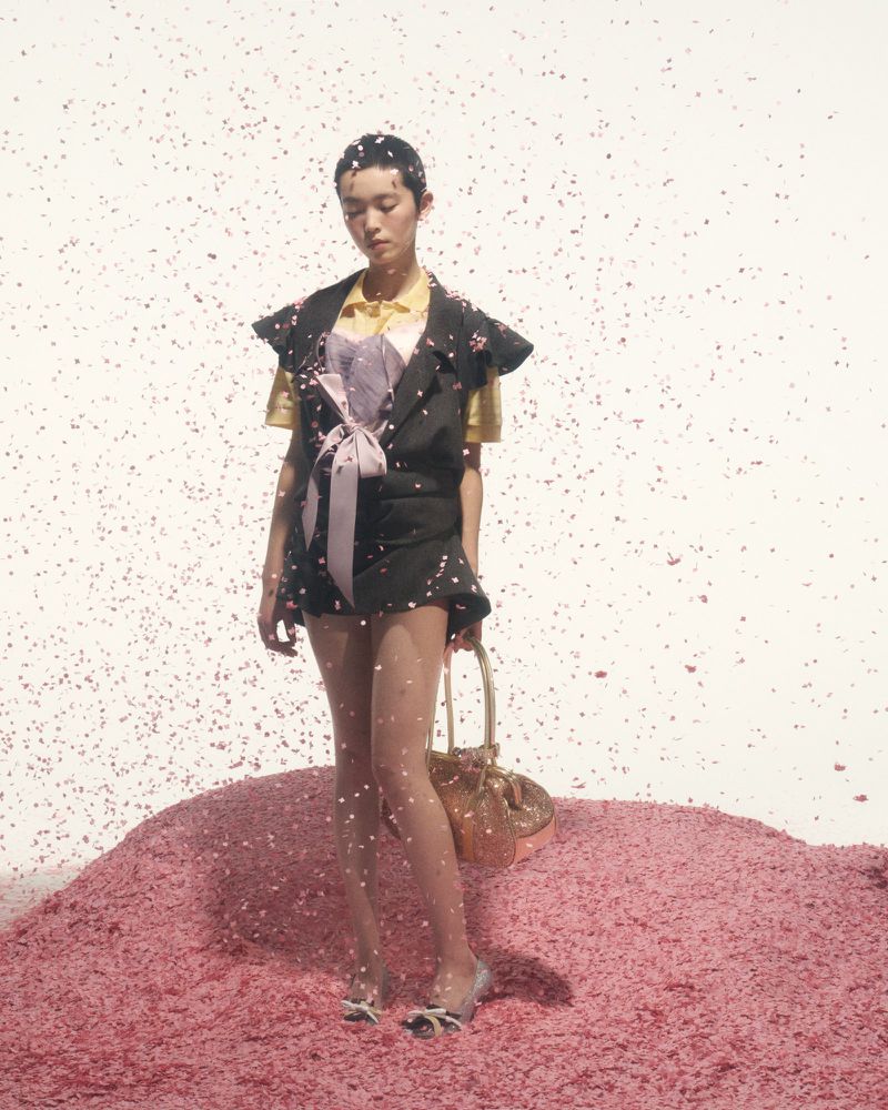 Asian woman poses on pink confetti pile with a gold purse and black suit in a studio setting with confetti falling all around her.