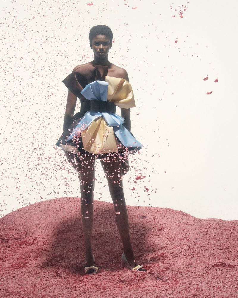 Dark-skinned female fashion model stands on pile of pink material, wearing short, modern dress against white background. Fashion shot.