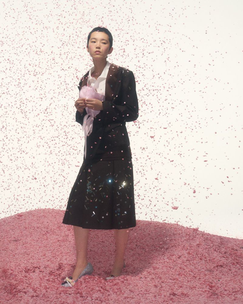 Asian female model in blazer and skirt stands in surreal confetti scene, holding pink prop and wearing glitter heels in photo studio.