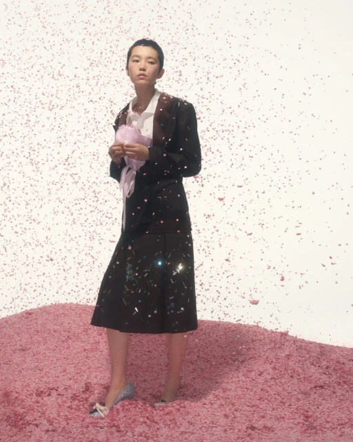 Asian female model in blazer and skirt stands in surreal confetti scene, holding pink prop and wearing glitter heels in photo studio.