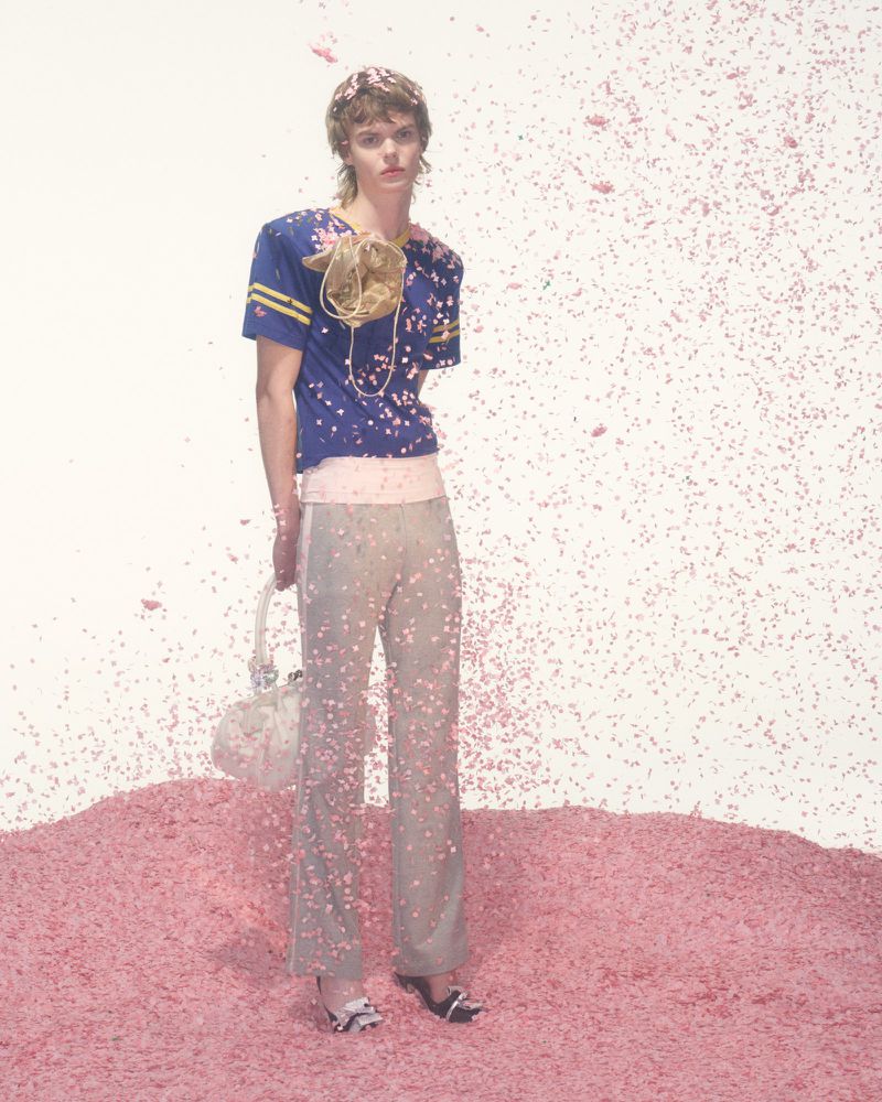 Fashion shot of an adult model in a blue and yellow top and gray pants, standing on a pink surface with confetti against a white backdrop.