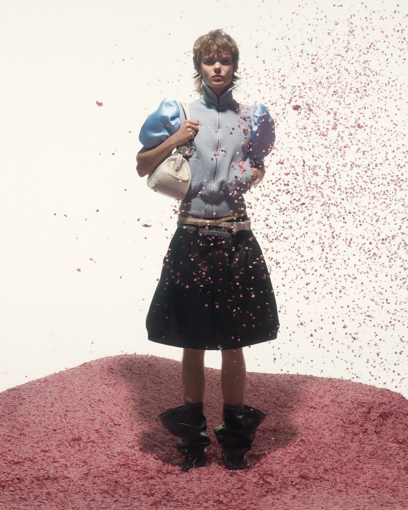 Fashion shot of a model in puffy-sleeved top, skirt, and large shoes standing on a pink pile surrounded by pink particles in studio.