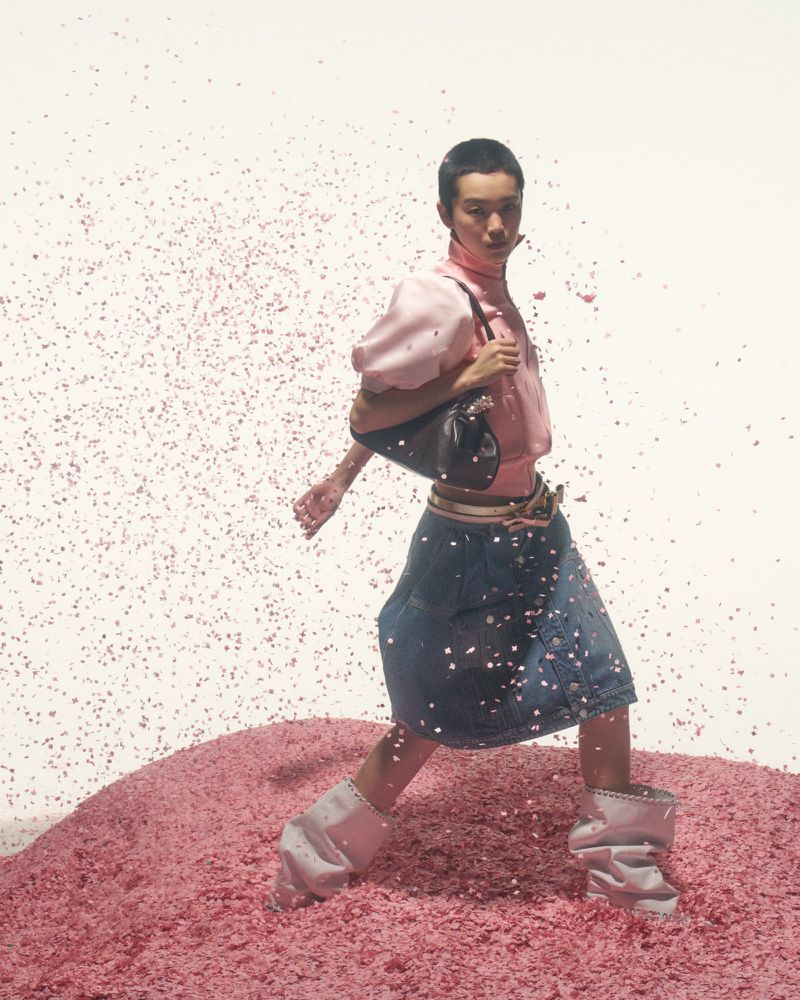 Fashion shot of an Asian woman in pink blouse and denim skirt walking on pink glitter, holding a handbag, set against a white background.