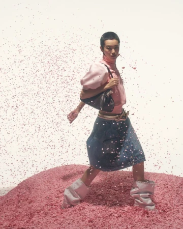 Fashion shot of an Asian woman in pink blouse and denim skirt walking on pink glitter, holding a handbag, set against a white background.