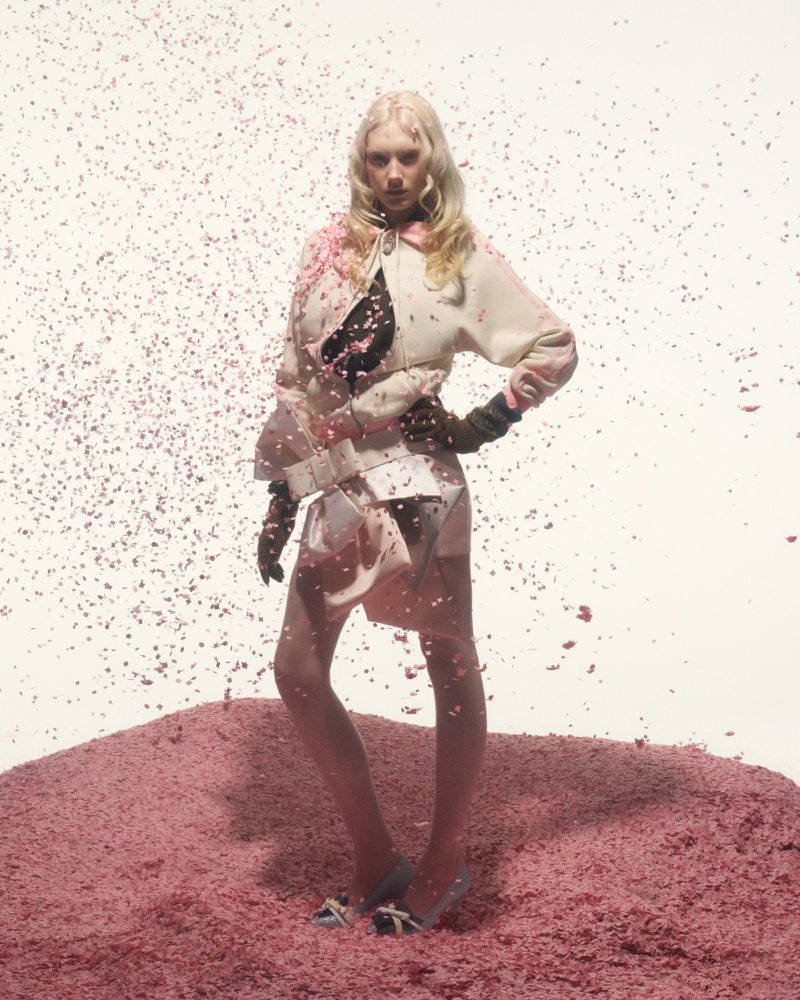 Fashion model in a pink and white jacket and skirt posing in a pink setting, surrounded by small pink confetti-like pieces in studio lighting.