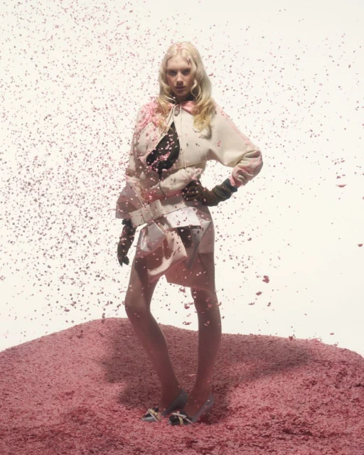 Fashion model in a pink and white jacket and skirt posing in a pink setting, surrounded by small pink confetti-like pieces in studio lighting.