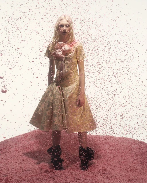 Fashion model poses in a golden dress amidst a pink backdrop and falling pink particles, creating a surreal and artistic fashion photograph.