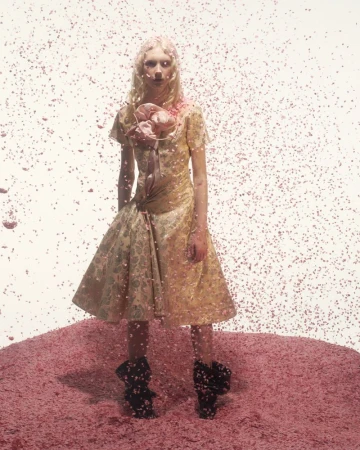Fashion model poses in a golden dress amidst a pink backdrop and falling pink particles, creating a surreal and artistic fashion photograph.