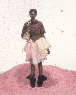 Model standing on pink mound in striped polo shirt and layered skirt holding handbag for August Barron