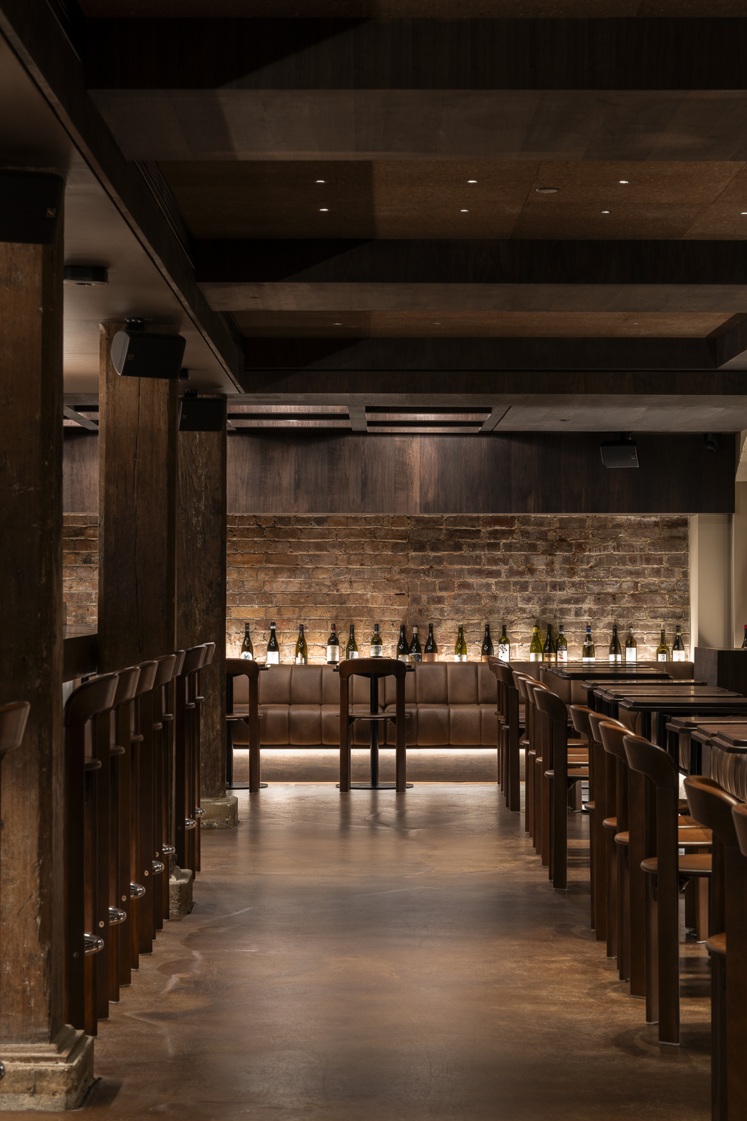 Interior view of a restaurant featuring wooden chairs and tables, exposed brick walls, and decorative wine bottles on a shelf.