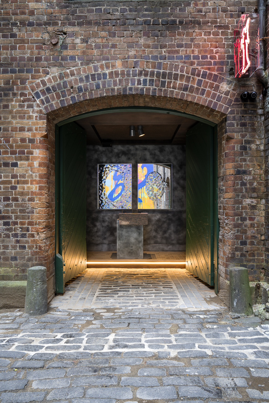 Urban art gallery entrance featuring colorful paintings inside a brick building with an arched doorway and cobblestone pavement outside.