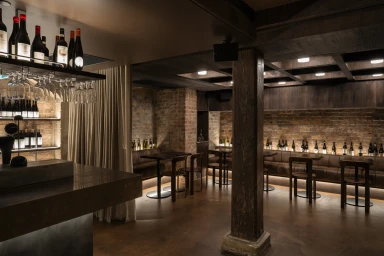Interior view of a moody bar with brick walls, leather booths, wooden tables, and bottles of wine on illuminated shelves.