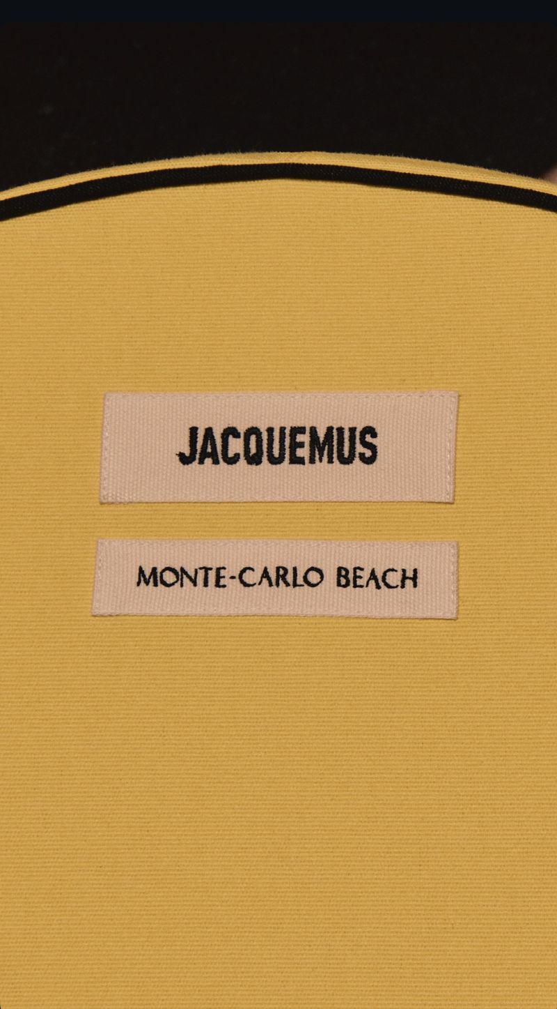 Close-up of a yellow fabric item featuring two labels with black text reading 'JACQUEMUS' and 'MONTE-CARLO BEACH' respectively.