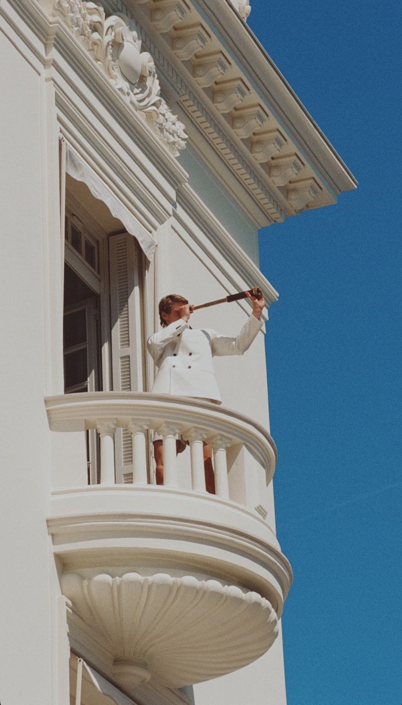 Adult male standing on a balcony looking through a spyglass with blue skies in the background and white building architecture.