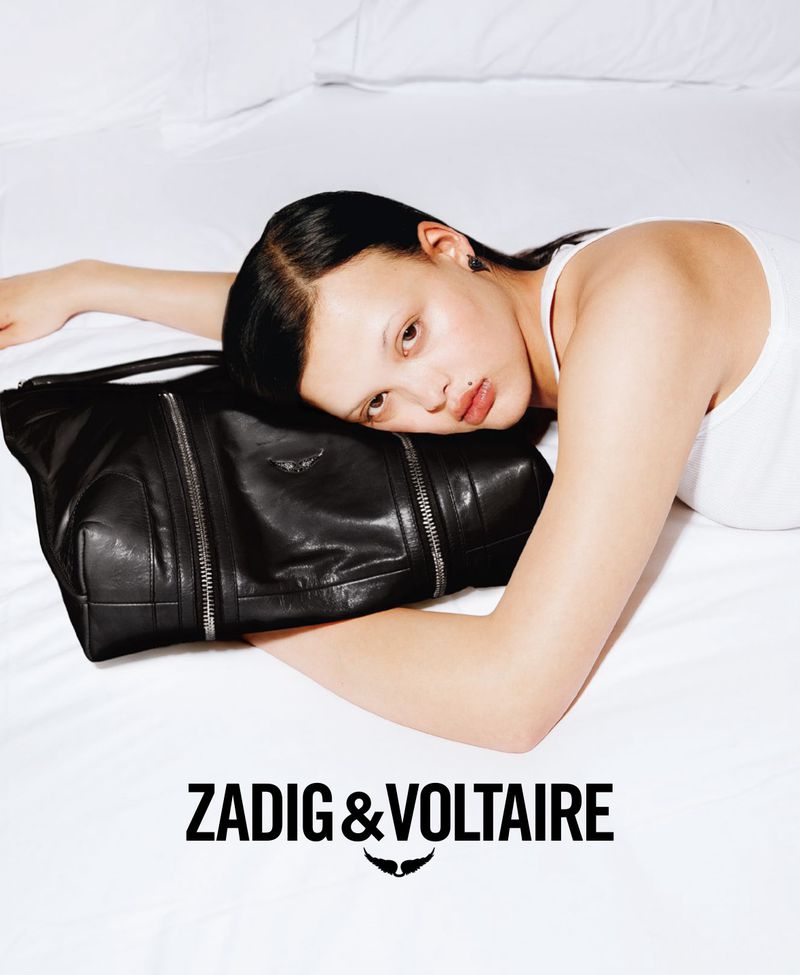 Fashion shot of a woman lying on a white sheet with a black leather handbag, with the Zadig & Voltaire logo at the bottom.
