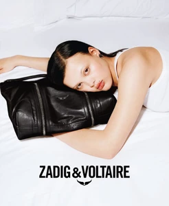 Model lying on white bedding resting her head against a large black leather handbag for Zadig & Voltaire