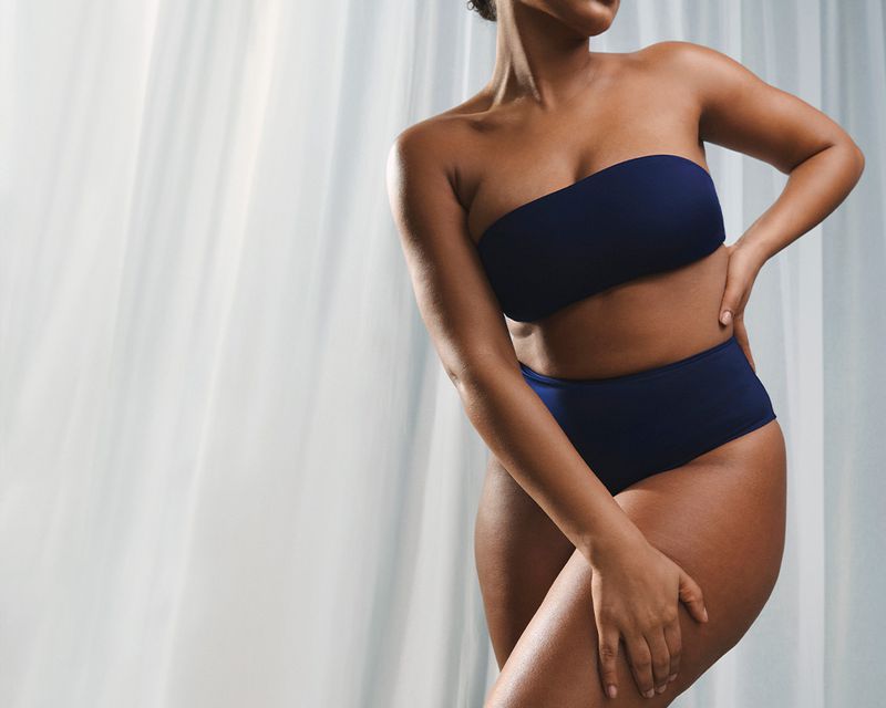 Cropped view of a woman with dark skin in a two-piece blue bathing suit against a bright white backdrop, posing indoors.