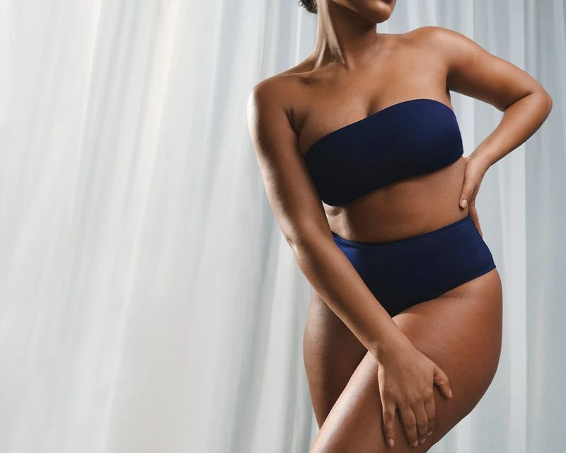Cropped view of a woman with dark skin in a two-piece blue bathing suit against a bright white backdrop, posing indoors.
