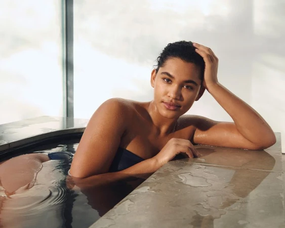 Woman relaxes in a bathtub by a window with natural light. She rests her head on her hand and looks toward the camera.