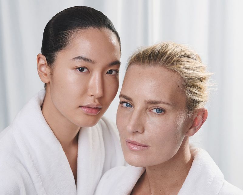 Two women in white bathrobes pose indoors, with soft lighting enhancing their natural skin tones in a beauty or skincare context.