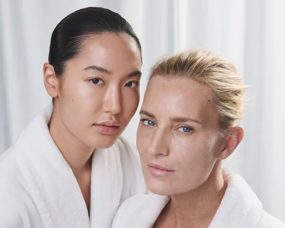 Two women in white bathrobes pose indoors, with soft lighting enhancing their natural skin tones in a beauty or skincare context.