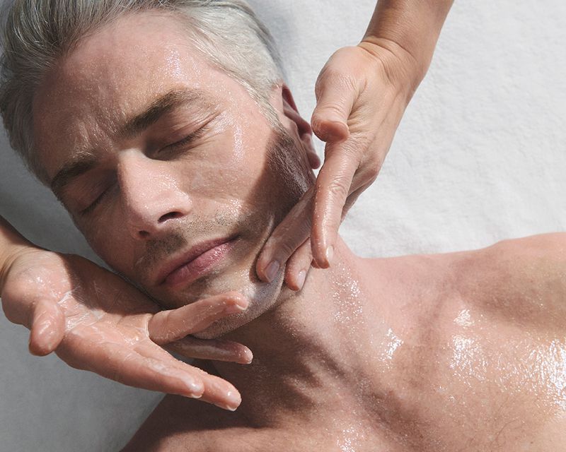 Adult man receives a facial massage. A pair of hands massages his face, neck, and chest. He has light skin and is lying down.