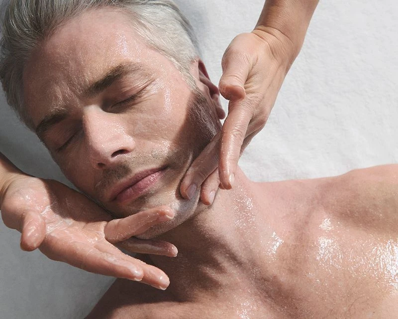 Adult man receives a facial massage. A pair of hands massages his face, neck, and chest. He has light skin and is lying down.