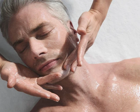 Adult man receives a facial massage. A pair of hands massages his face, neck, and chest. He has light skin and is lying down.