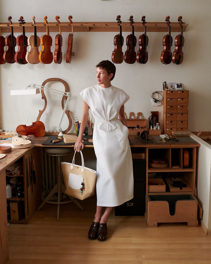 Woman in white dress stands in luthier's workshop. Violins hang on the wall. Natural light and vintage aesthetic enhance the scene.