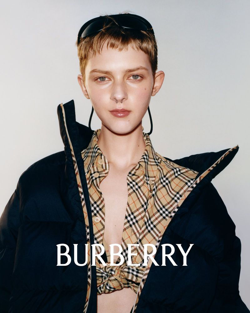 Young woman poses in a Burberry shirt and jacket against a neutral backdrop, wearing black sunglasses atop her short, light-brown hair.