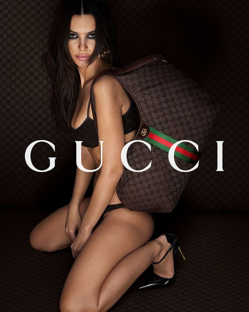 Woman in black lingerie poses with brown GG pattern Gucci bag against a matching patterned backdrop in a studio fashion shot.