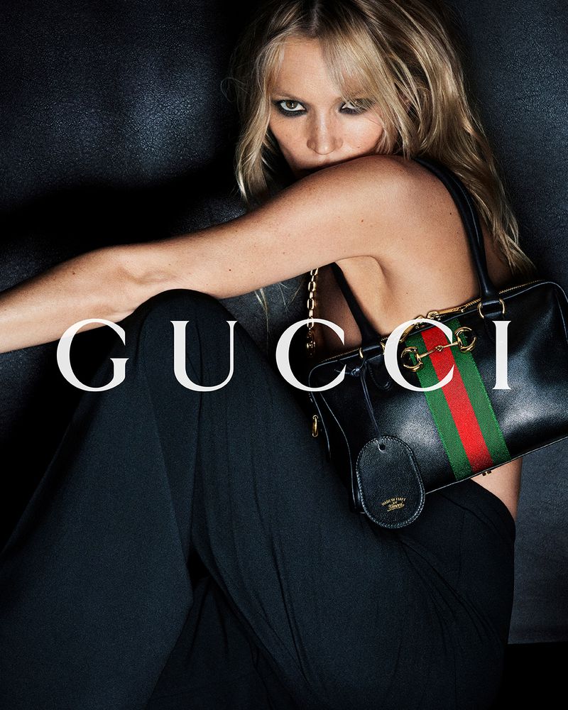 Fashion shot of a blonde adult female model with Gucci handbag in front of a textured dark grey background with Gucci logo.