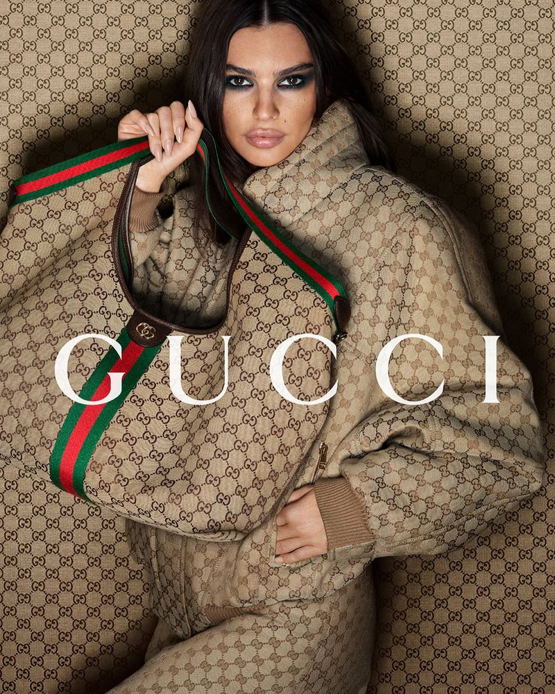 Emily Ratajkowski posing in a monogram Gucci puffer jacket holding a matching Gucci handbag against a branded fabric background