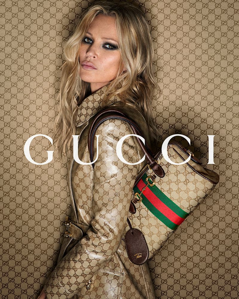 Blonde woman, Kate Moss, poses wearing a Gucci coat and holding a Gucci bag in front of a Gucci logo wallpaper background.