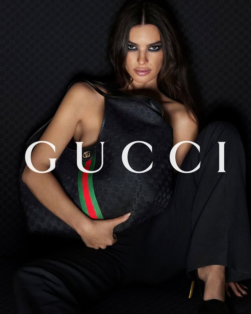 A woman models a Gucci handbag in a studio setting. The handbag features a green and red stripe with the Gucci logo in white text.