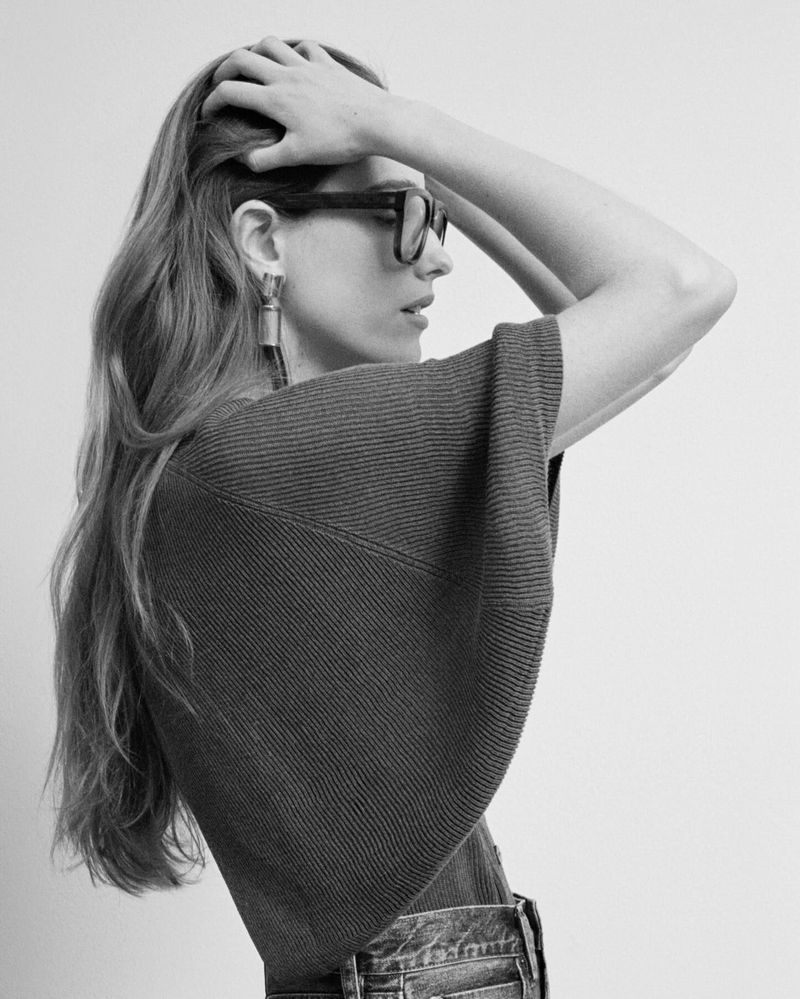 Black and white shot of a woman with long wavy hair wearing glasses, an earring, and a knit top against a neutral background.