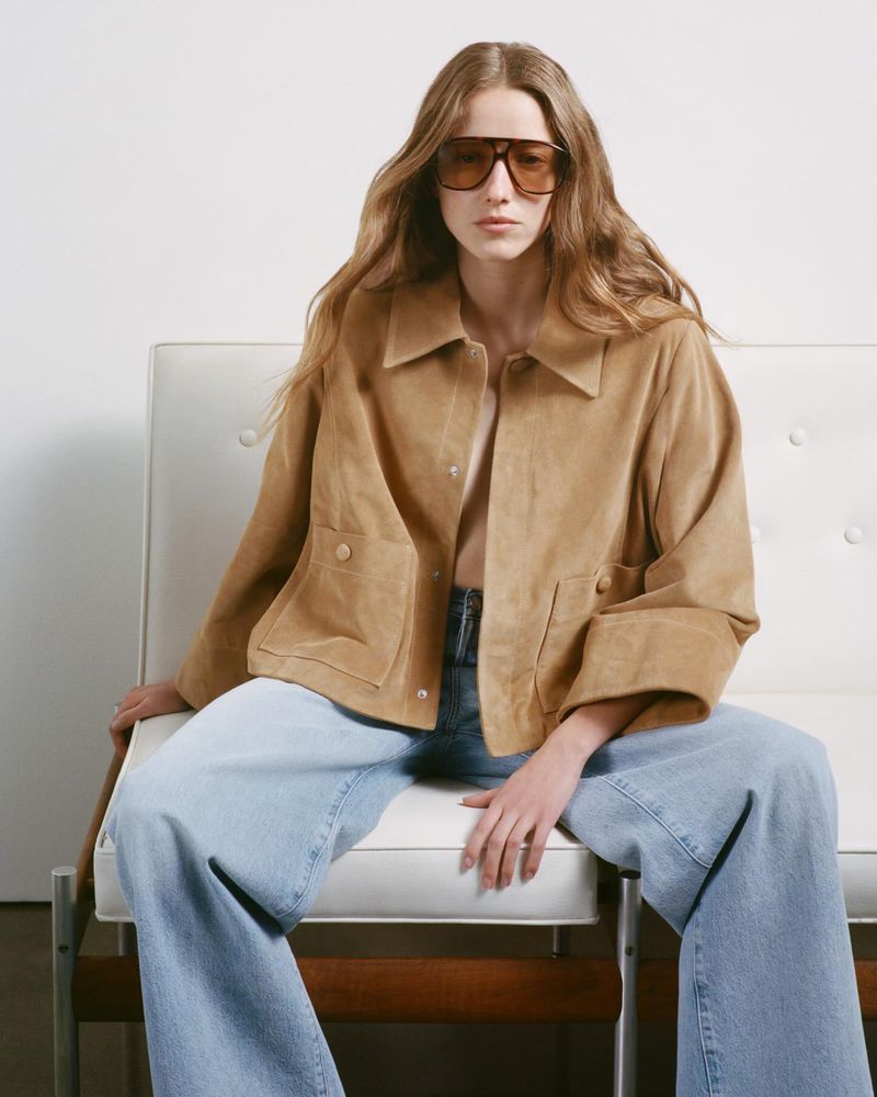 A blond woman poses on a white chair in a tan suede jacket and light blue jeans against a white background in a fashion shot.
