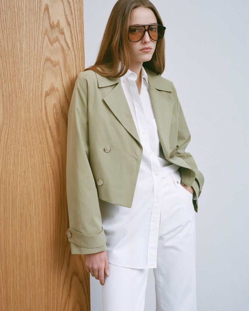 A woman with sunglasses stands in front of a wooden panel, wearing a cropped light-green jacket, white button-down shirt, and white pants.