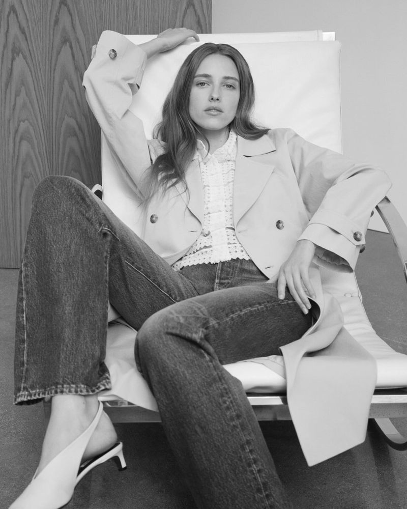 A woman in a beige jacket and blue jeans poses on a white chair, with a neutral background, showcasing a casual yet stylish fashion look.